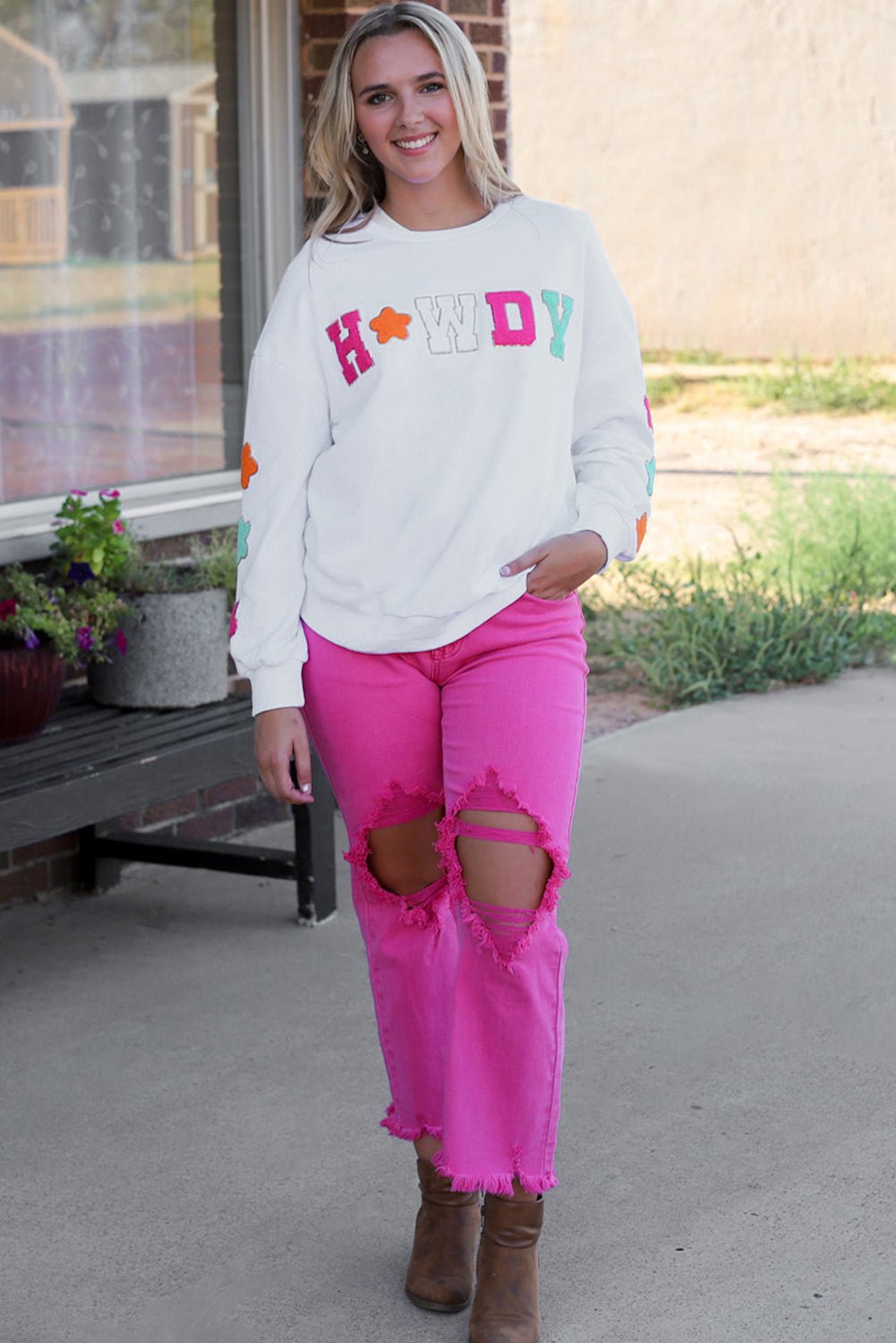 Woman wearing Threaded Pear Howdy patch sweatshirt in white with colorful letters paired with pink ripped jeans and brown boots